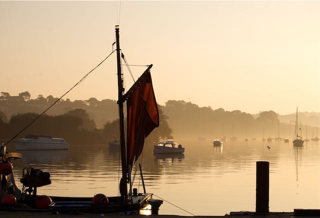 Early morning spring mist over water with trees in the background. A boat with a mizen mast front and centre. Beautiful morning light of amber and sharp relief shadows three minutes walk from the cottage.